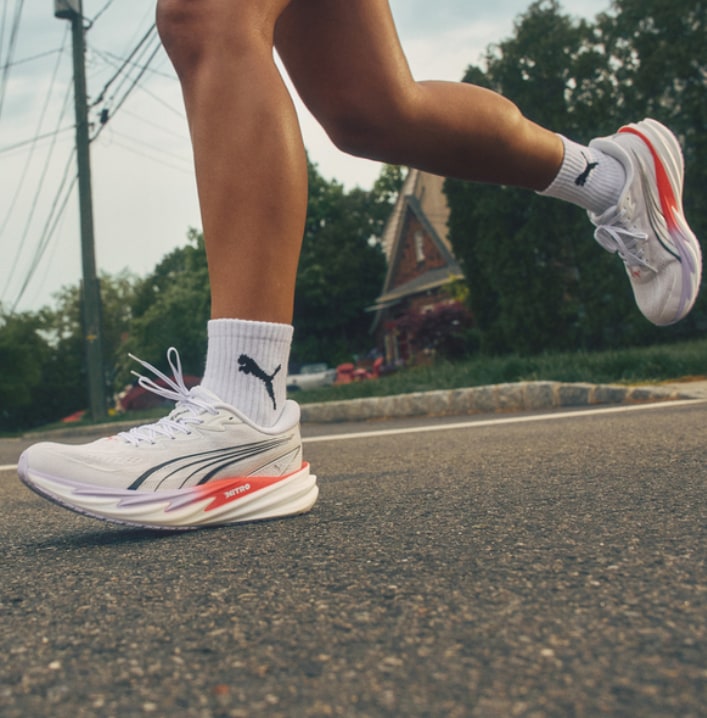 A person's legs and feet running on an asphalt road, wearing white athletic shoes with pink soles and white socks.